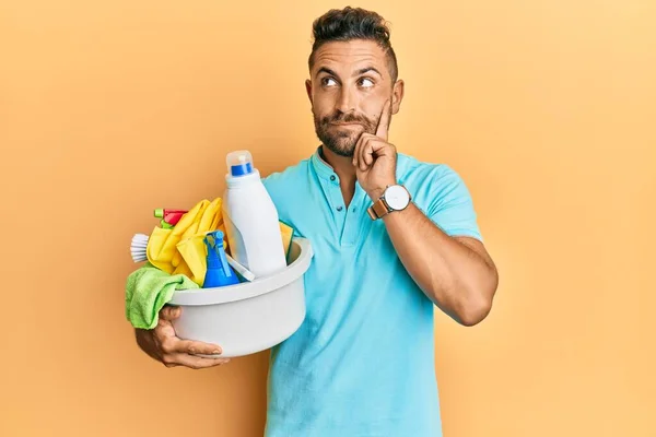 Handsome man with beard holding cleaning products serious face thinking about question with hand on chin, thoughtful about confusing idea