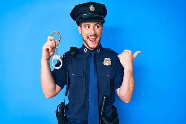 Young caucasian man wearing police uniform holding handcuffs pointing thumb up to the side smiling happy with open mouth