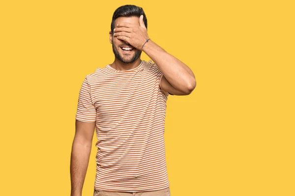 カジュアルな服を着たハンサムなヒスパニック系の男が笑顔で 驚きのために目を覆う顔で手で笑っています 視覚障害の概念 — ストック写真