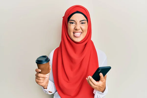 若いです美しいですヒスパニック系の女の子身に着けていますイスラムヒジャーブ飲むAコーヒーUsignスマートフォンステッキング舌アウト幸せなで面白い式 — ストック写真