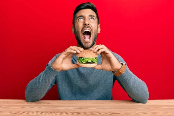 Bonito Homem Hispânico Comendo Saboroso Hambúrguer Clássico Irritado Louco Gritando — Fotografia de Stock