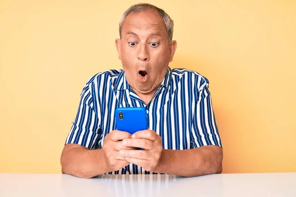 Senior handsome man with gray hair using smartphone sitting on the table scared and amazed with open mouth for surprise, disbelief face