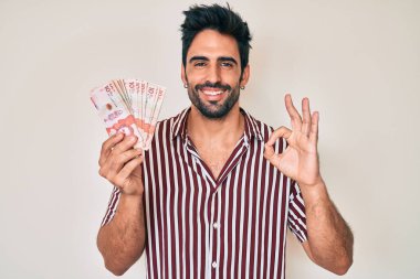 Sakallı yakışıklı İspanyol adam Kolombiya pezosu tutuyor. Parmaklarıyla imza atıyor, gülümseyerek mükemmel bir sembol oluşturuyor. 