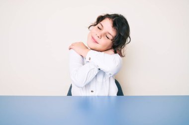 Tatlı İspanyol çocuk gündelik giysiler giyip masanın üzerinde oturmuş kendine sarılıyor mutlu ve pozitif, kendinden emin gülümsüyor. Sevgi ve özgüven. 