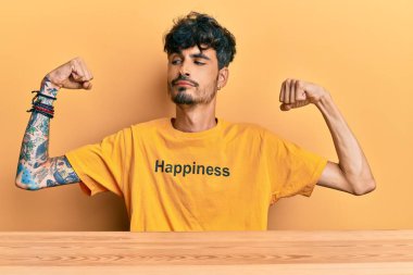 Üzerinde mutluluk mesajı olan tişört giyen genç İspanyol adam masaya oturmuş gururla gülümseyen kol kaslarını gösteriyor. fitness konsepti. 