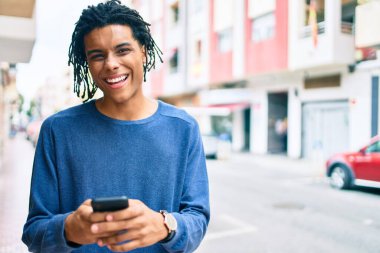 Genç Afrikalı Amerikalı adam şehrin caddesinde akıllı telefon kullanarak mutlu gülümsüyor..