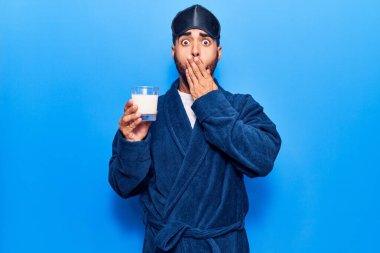 Uyku maskesi takan ve bornoz giyen, ağzı ağızla kapalı süt içen, şok olmuş ve hata yapmaktan korkan genç bir İspanyol. şaşırmış ifade 