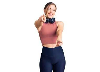 Güzel beyaz kadın spor kıyafetleri giyiyor ve kulaklık takıyor parmaklarıyla seni ve kamerayı işaret ediyor, gülümsüyor ve neşeli. 