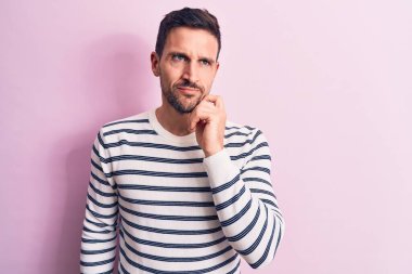 Sıradan çizgili süveter giyen yakışıklı genç bir adam pembe arka planda dikilirken parmakları çenesinde şüpheye odaklanmış ve yukarı bakıp merak ediyordu.