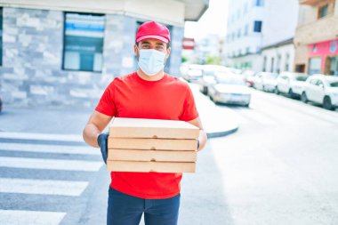 Üniforma giyen ve koronavirüs koruma maskesi takan genç teslimatçı. Kasaba caddesinde pizza kutularını teslim ediyorum..