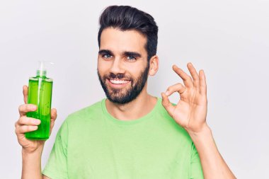 Sakallı genç yakışıklı adam aloe vera krem şişesi giyiyor tamam işareti yapıyor parmaklarıyla gülümseyerek mükemmel bir sembol oluşturuyor. 