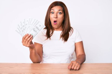 Orta yaşlı esmer kadın elinde dolarlarla masada oturuyor. Korkmuş ve şaşkın bir şekilde ağzı açık bekliyor. 