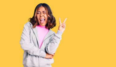 Genç, güzel, melez, spor kıyafetleri giyen, kameraya gülümseyen ve zafer işareti yapan bir kadın. İki numara.. 