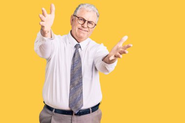 İş kıyafetleri giymiş yaşlı, kır saçlı bir adam kameraya sarılıp gülümsüyor. Mutluluğu kucaklayan neşeli bir ifade.. 