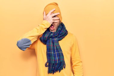 Kışlık giysiler giymiş genç, yakışıklı kel bir adam. Şoka girmiş, yüzünü ve gözlerini kapamış, parmaklarından korkuyla bakıyor. 