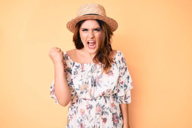 Yazlık elbise giyen, şapkalı, kızgın ve kızgın genç beyaz bir kadın öfkeyle bağırırken yumruğunu kaldırıyor. öfke ve agresif konsept. 