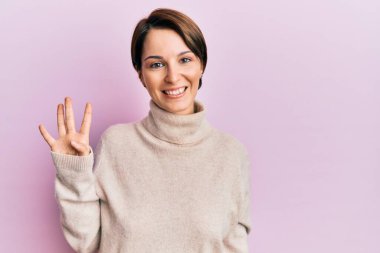 Kısa saçlı esmer kadın, günlük kışlık süveter giyiyor ve dört numaralı parmaklarıyla işaret ediyor. Kendinden emin ve mutlu gülümsüyor.. 