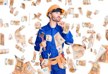 Elektirikçi üniforması giyen genç İspanyol adam İngiliz anahtarını elinde tutarak ciddi bir yüz ifadesiyle eli çenesinde soru sormayı düşünüyor, kafa karıştırıcı bir fikir hakkında düşünceli.