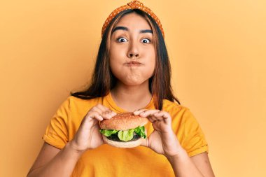 Lezzetli bir burger yiyen genç Latin kadın komik yüzlü yanakları şişiriyor. Ağzı havayla şişer, havayı yakalar.. 