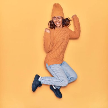 Yün şapka ve süveter giyen, mutlu gülümseyen genç, güzel Latin bir kadın. Yüzünde tebessümle zıplıyor, sarı arka planda yumruklar havada kutluyor.