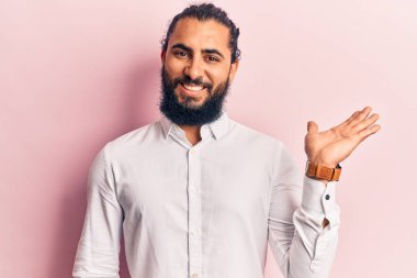 Gündelik giysiler giyen genç Arap adam neşeli bir şekilde gülümsüyor ve avuç içi kameraya bakıyor.. 