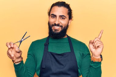 Genç Arap adam berber önlüğü takıyor gülümsüyor mutlu bir şekilde eli ve parmağı yana doğru işaret ediyor 