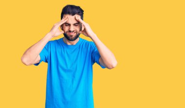 Sakallı, gündelik tişörtlü, baş ağrısı çeken, çaresiz ve stresli genç bir adam. Ağrı ve migren yüzünden. Eller başının üstünde.. 