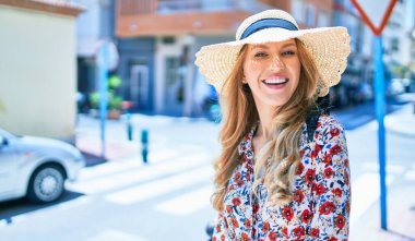 Yaz şapkası takmış, mutlu bir şekilde gülümseyen güzel sarışın bir kadın. Şehrin caddesinde yüzünde gülümsemeyle dikilirken.