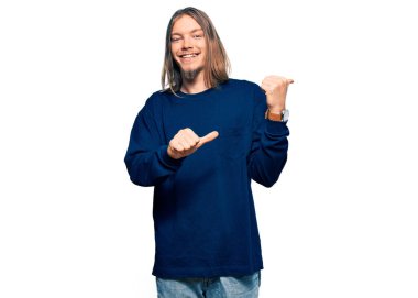 Uzun saçlı yakışıklı beyaz adam, günlük kışlık süveter giyiyor. Arka tarafı işaret ediyor. El ve baş parmakları havada, kendinden emin. 