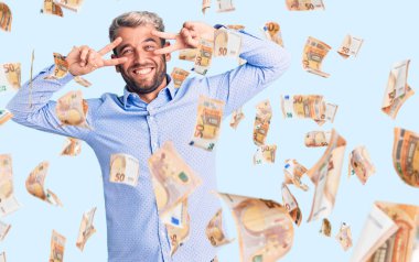 Zarif tişört giyen yakışıklı genç adam barış sembolü yapıyor. Yüzünde parmaklar, gülümseyerek zaferi gösteriyor.