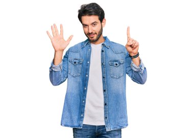 Latin asıllı genç adam gündelik giysiler giyip altı numaralı parmaklarıyla işaret ederken kendinden emin ve mutlu bir şekilde gülümsüyor.. 