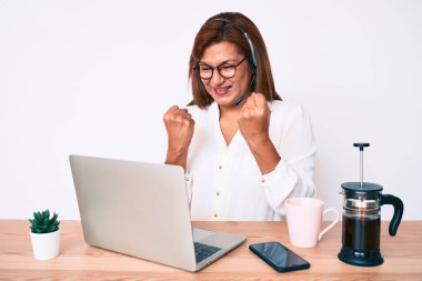 Orta yaşlı esmer kadın ofiste çalışan operatör kulaklığı takıyor. Kolları havada ve gözleri kapalı bir şekilde başarıyı kutluyor ve şaşırıyor. 