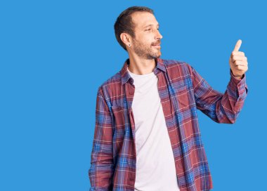 Gündelik kıyafetler giyen, gururlu, gülümseyen, baş parmak kaldıran genç yakışıklı bir adam. 