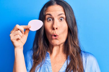 Genç, güzel, esmer bir kadın mavi arka planda sünger kullanarak makyaj yapıyor. Şaşırmak için ağzı açık ve korkmuş, inançsız bir yüz.
