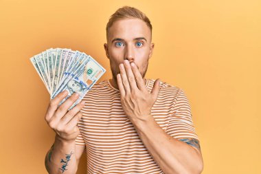 Dolarları tutan genç beyaz bir adam ağızlarını eliyle kapatıyor, şaşırmış ve hata yapmaktan korkuyor. şaşırmış ifade 