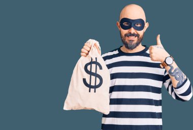 Hırsız maskesi takan yakışıklı bir genç dolar sembollü para çantası tutuyor mutlu ve olumlu gülümsüyor, başparmağıyla mükemmel ve onay işareti yapıyor. 
