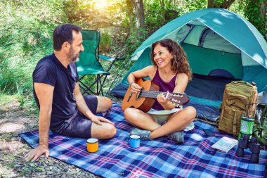 Ormanda kamp yaparken gülümseyen orta yaşlı bir çift. Yerde oturup klasik gitar çalarak şarkı söylemek.