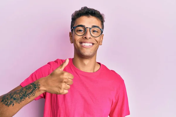 年轻英俊的非洲裔美国男人戴着粉色背景的眼镜 手举着快乐的大拇指 赞成的表情看着相机显示的成功 — 图库照片