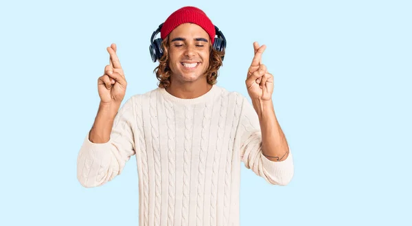 Young Hispanic Man Listening Music Using Headphones Gesturing Finger Crossed — Stock Photo, Image