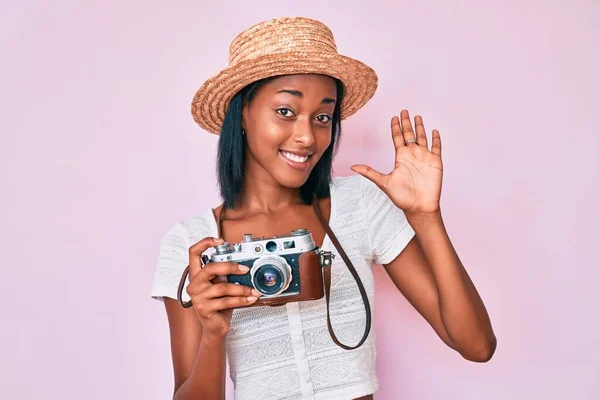 Young african american woman wearing summer hat holding vintage camera waiving saying hello happy and smiling, friendly welcome gesture