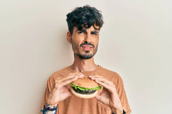 Jovem Hispânico Comendo Saboroso Clássico Hambúrguer Sem Noção Expressão Confusa — Fotografia de Stock