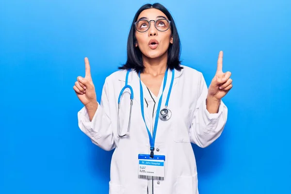 Young beautiful latin woman wearing doctor stethoscope and id card amazed and surprised looking up and pointing with fingers and raised arms.