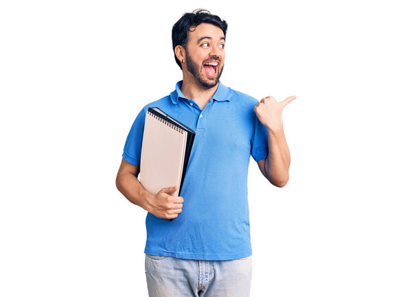 Young hispanic man holding notebook pointing thumb up to the side smiling happy with open mouth 