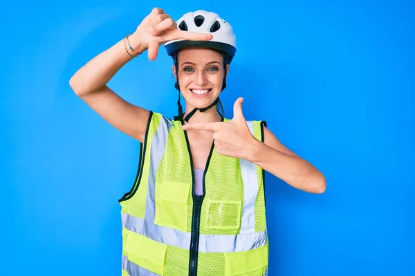 Young caucasian girl wearing bike helmet and reflective vest smiling making frame with hands and fingers with happy face. creativity and photography concept.