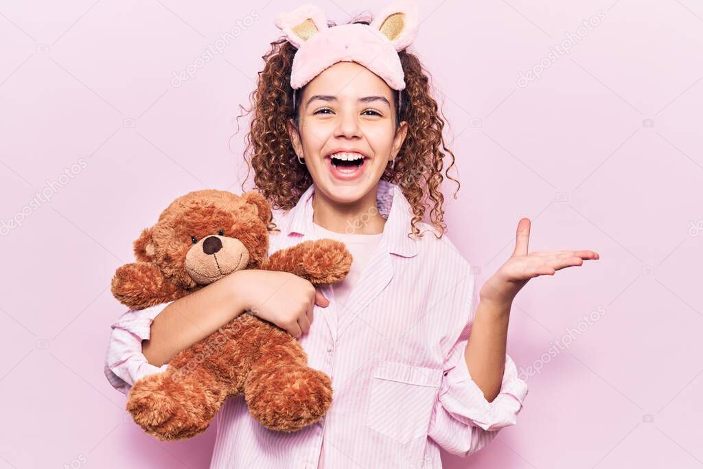 Beautiful kid girl with curly hair wearing sleep mask and pajamas holding teddy bear celebrating victory with happy smile and winner expression with raised hands 