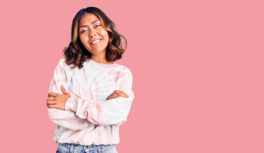 Genç, güzel, melez bir kadın, günlük kravat boyası kazak giyiyor, mutlu bir yüz, kollarını kavuşturup kameraya bakıyor. pozitif kişi. 