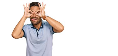 Sakallı, sıradan kıyafetler giyen, dürbün gibi hareket eden, dilini dışarı çıkaran, parmaklarından bakan yakışıklı bir adam. çılgın ifade. 