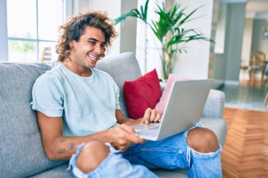 Evde dizüstü bilgisayar kullanan, mutlu gülümseyen genç İspanyol adam.