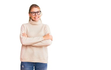 Balıkçı yaka kazak giyen ve kollarını kavuşturup kameraya bakan mutlu gözlüklü güzel sarışın kadın. pozitif kişi. 