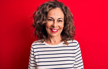 Orta yaşlı, esmer, çizgili tişört giyen güzel bir kadın kırmızı arka planda mutlu ve havalı bir gülümsemeyle dikiliyor. Şanslı kişi..